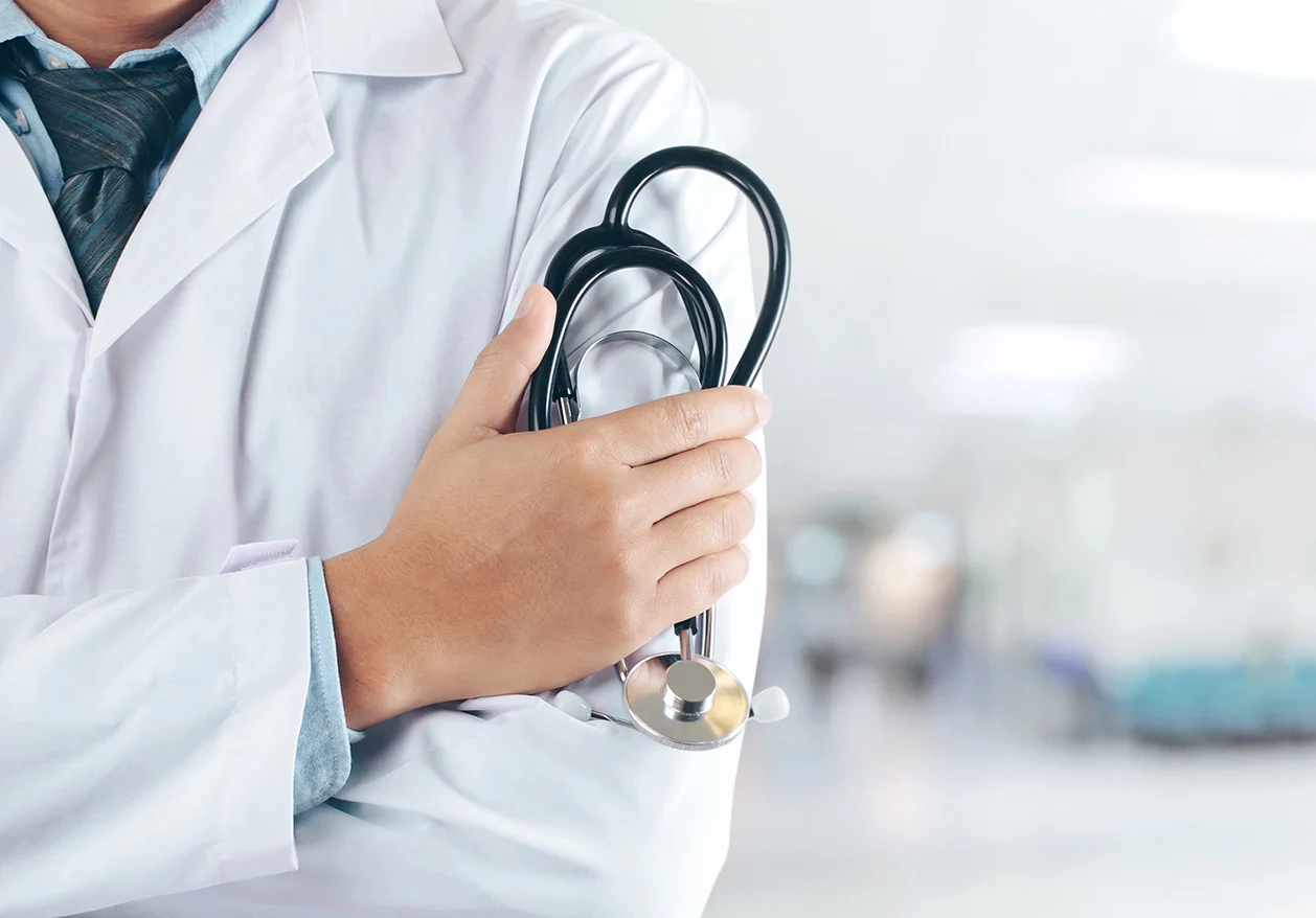 A male doctor in a lab coat holds a stethoscope and crosses his arms over his chest.